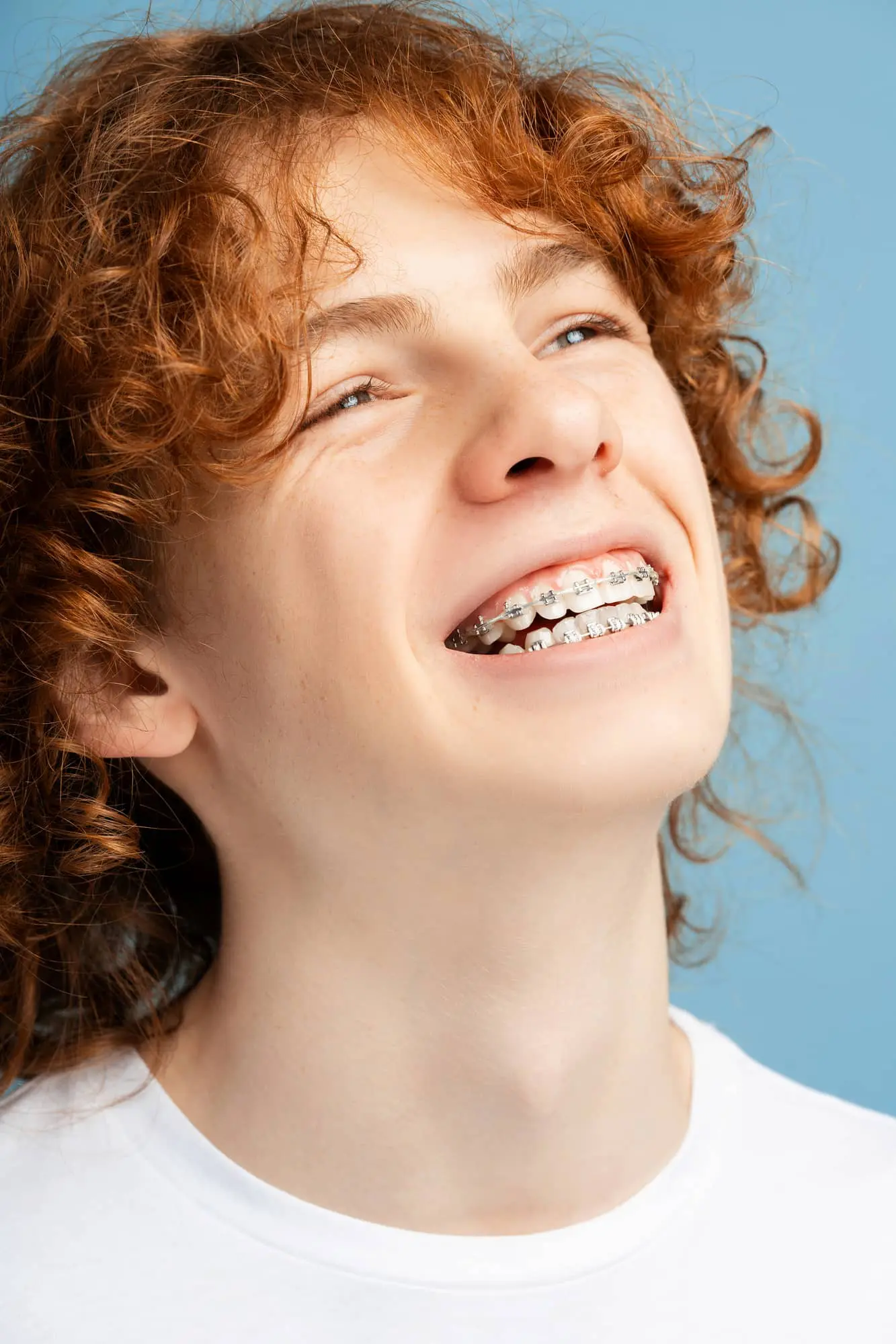 Person with curly red hair smiling, showing metal braces on teeth, wearing a white shirt against a blue background.