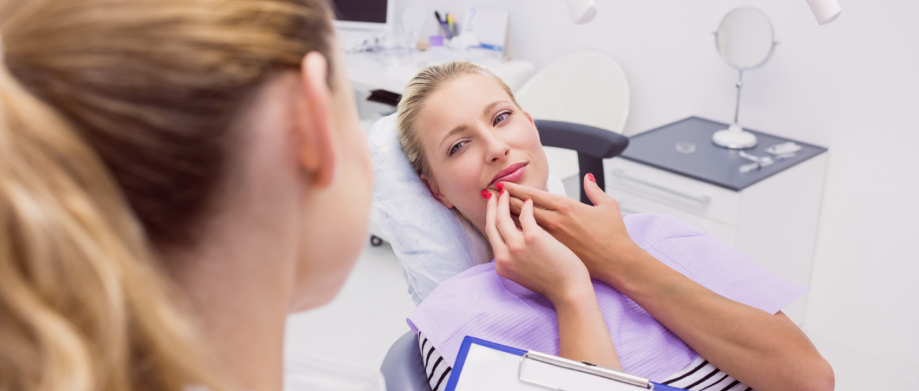 A woman is sitting in a dental chair touching her mouth. Another person, possibly a dentist, is holding a clipboard and facing her in an office setting.