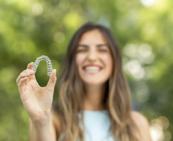 Person holding a clear orthodontic aligner with a blurred smile in the background and greenery around.
