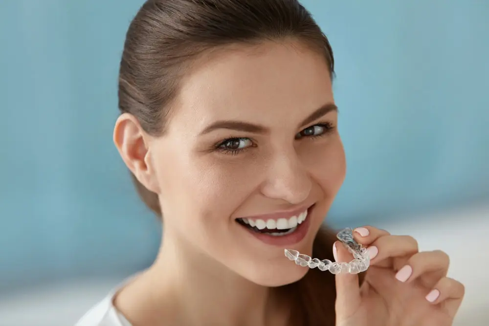 A woman smiles while holding a clear dental aligner near her mouth against a blue background, showcasing the results of the best orthodontics.