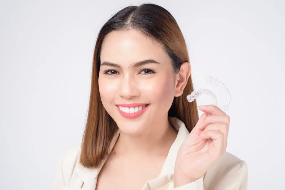 A woman smiling and holding a clear dental aligner near her face, wearing a cream-colored jacket, with a plain white background.