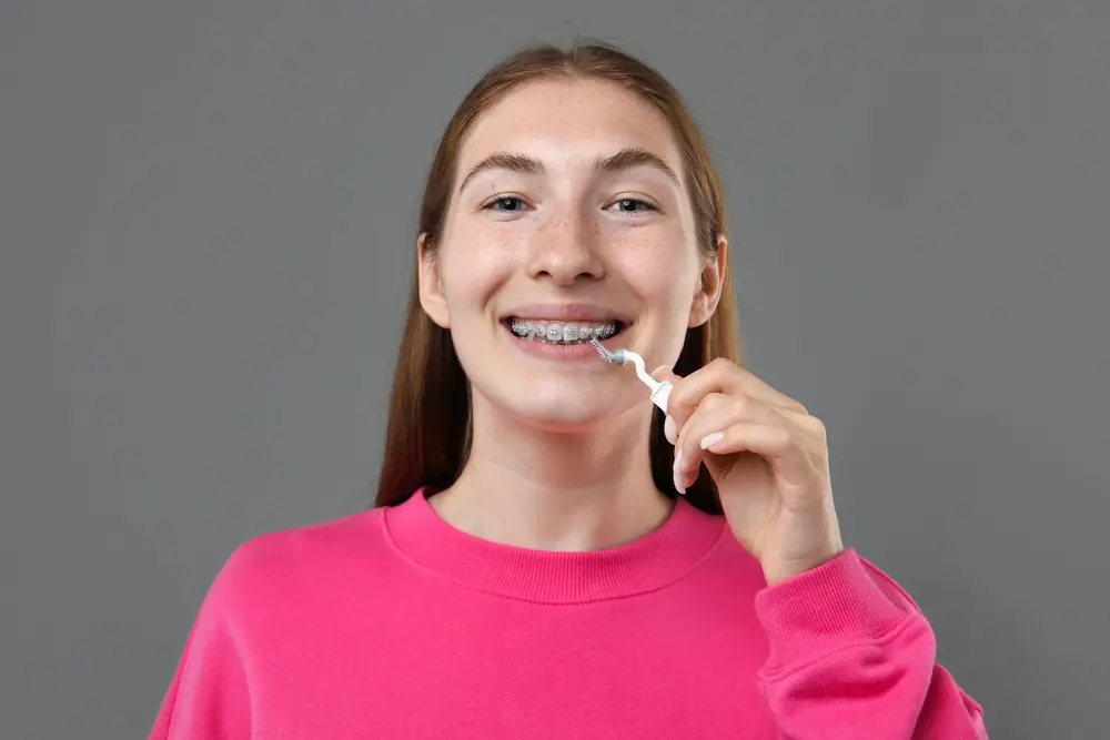 Young person with long hair in a pink sweater cleaning braces with a dental tool against a gray background