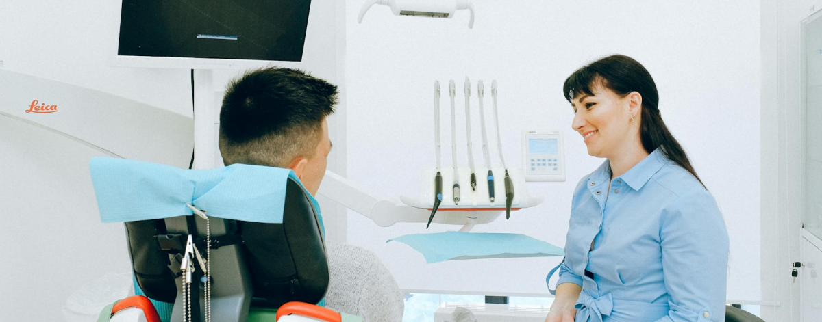 A dentist in a blue coat talks to a seated patient in a dental office with equipment visible in the background.