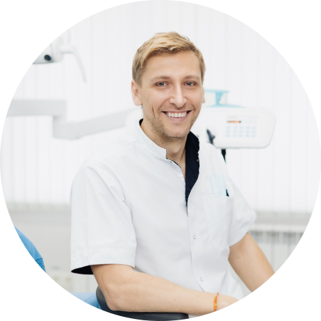 A dentist in a white coat smiles while seated in a dental office with equipment in the background.