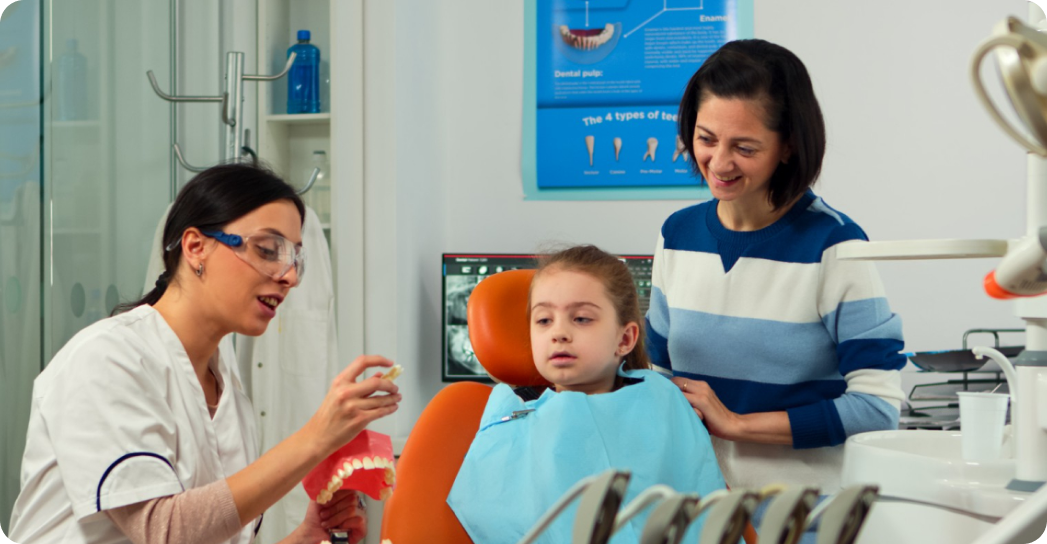 A dentist shows a dental model to a young girl sitting in a chair, while an adult stands beside them.