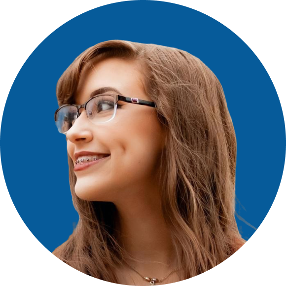 A person with long brown hair, glasses and braces smiles while looking to the side against a blue background.