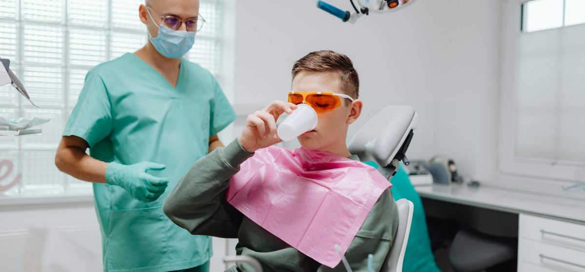 A dental patient wearing orange glasses drinks from a cup while seated in a dentist's chair. A dentist in scrubs and a mask stands nearby.