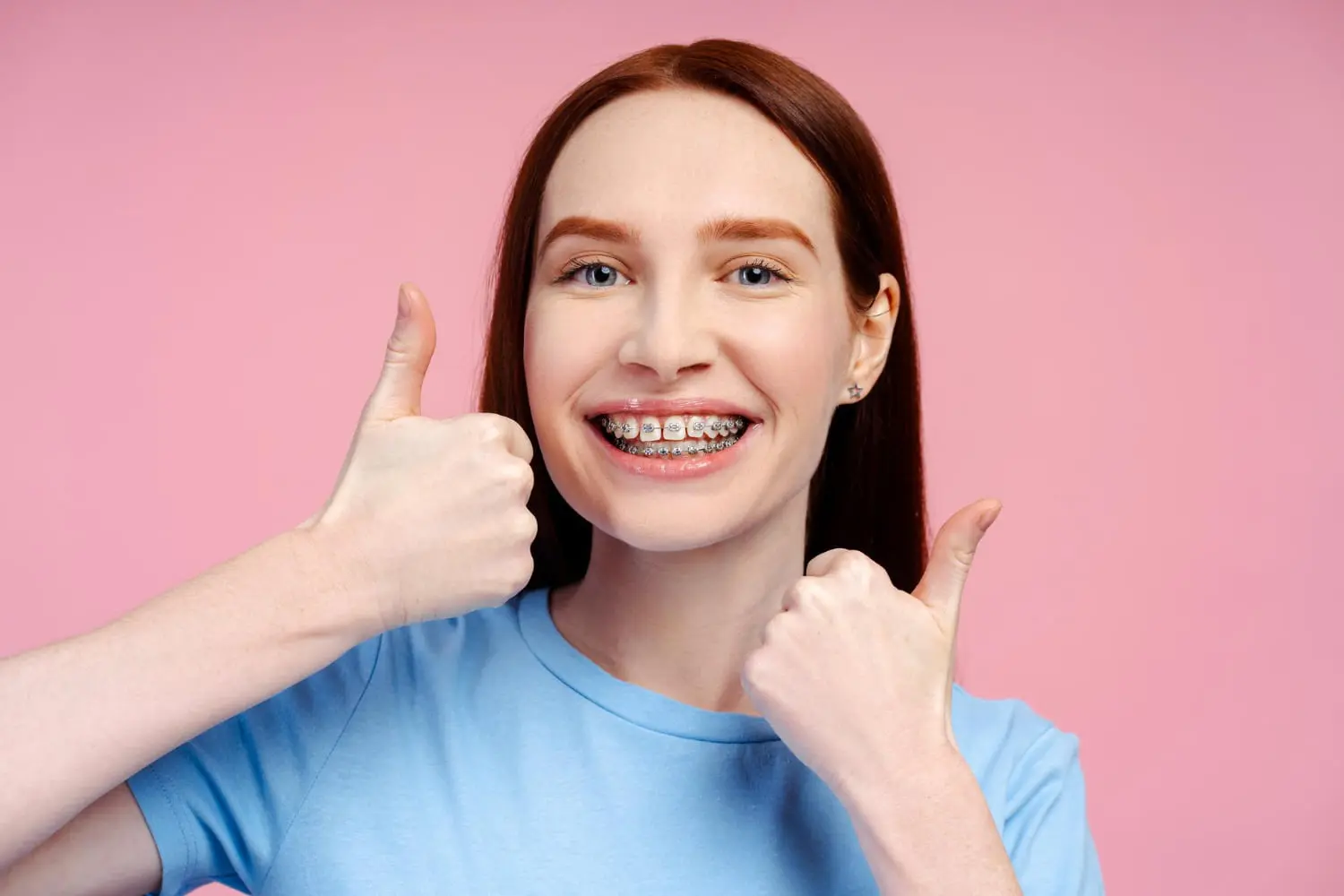 A smiling person with braces from Best Orthodontics gives two thumbs up, proudly wearing a light blue shirt against a vibrant pink background.