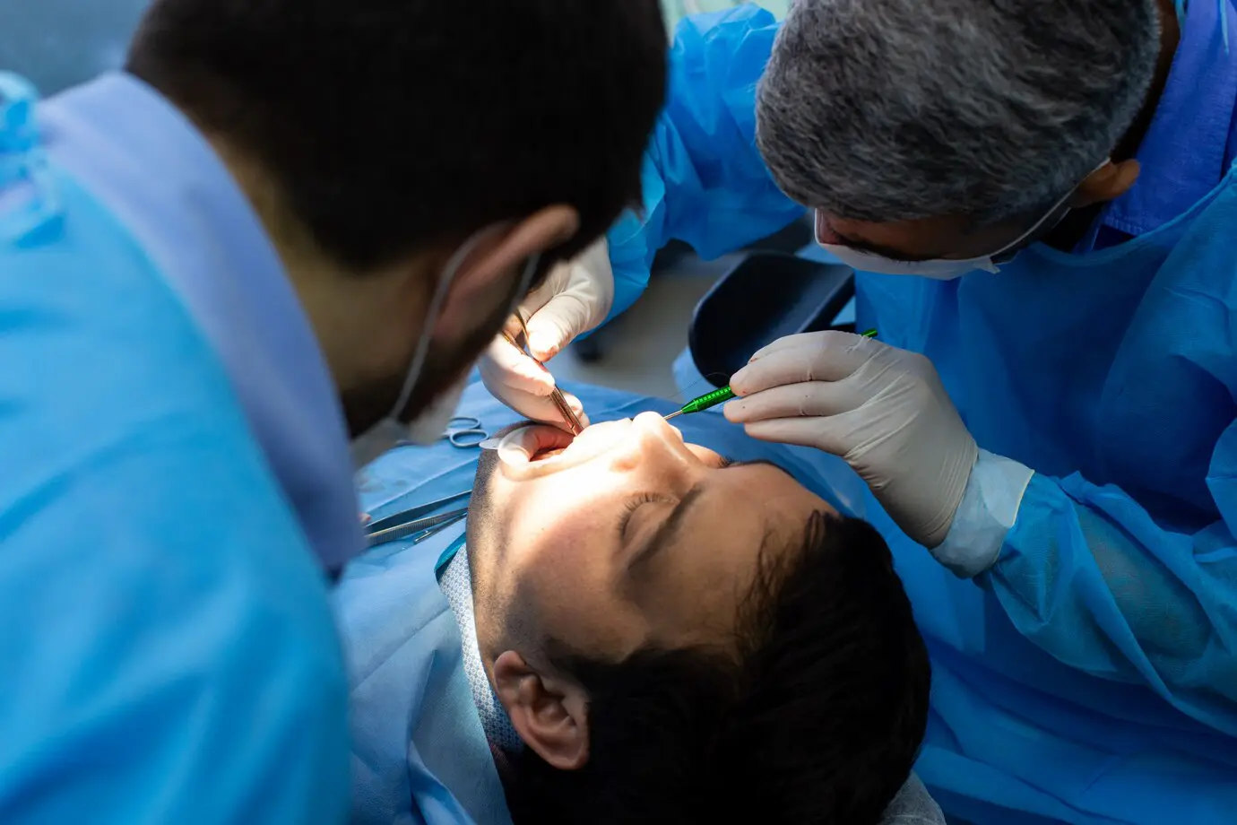 Two dentists in blue scrubs perform a dental procedure on a patient lying on an examination chair.