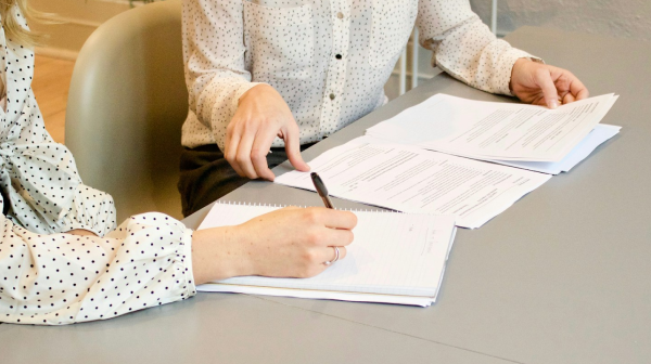 Two people sit at a table reviewing documents, with one person taking notes in a notebook.