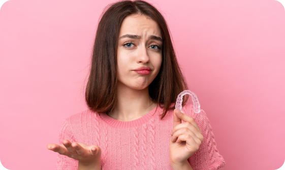 A woman in a pink sweater holds a clear dental aligner in one hand, while gesturing with the other against a pink background.