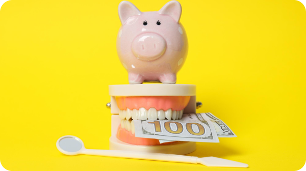 A piggy bank sits atop a model mouth holding a $100 bill, with a dental mirror and tool nearby, all against a yellow background.