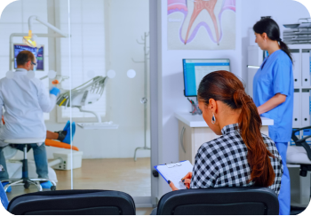 A woman fills out a form in a waiting area while a dentist attends to a patient in the examination room. A nurse works at a computer nearby.
