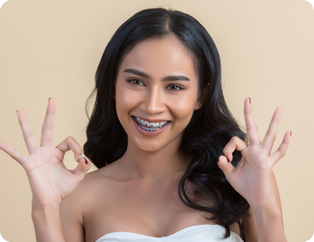 Person with long hair and braces smiling and making "OK" gestures with both hands against a beige background.