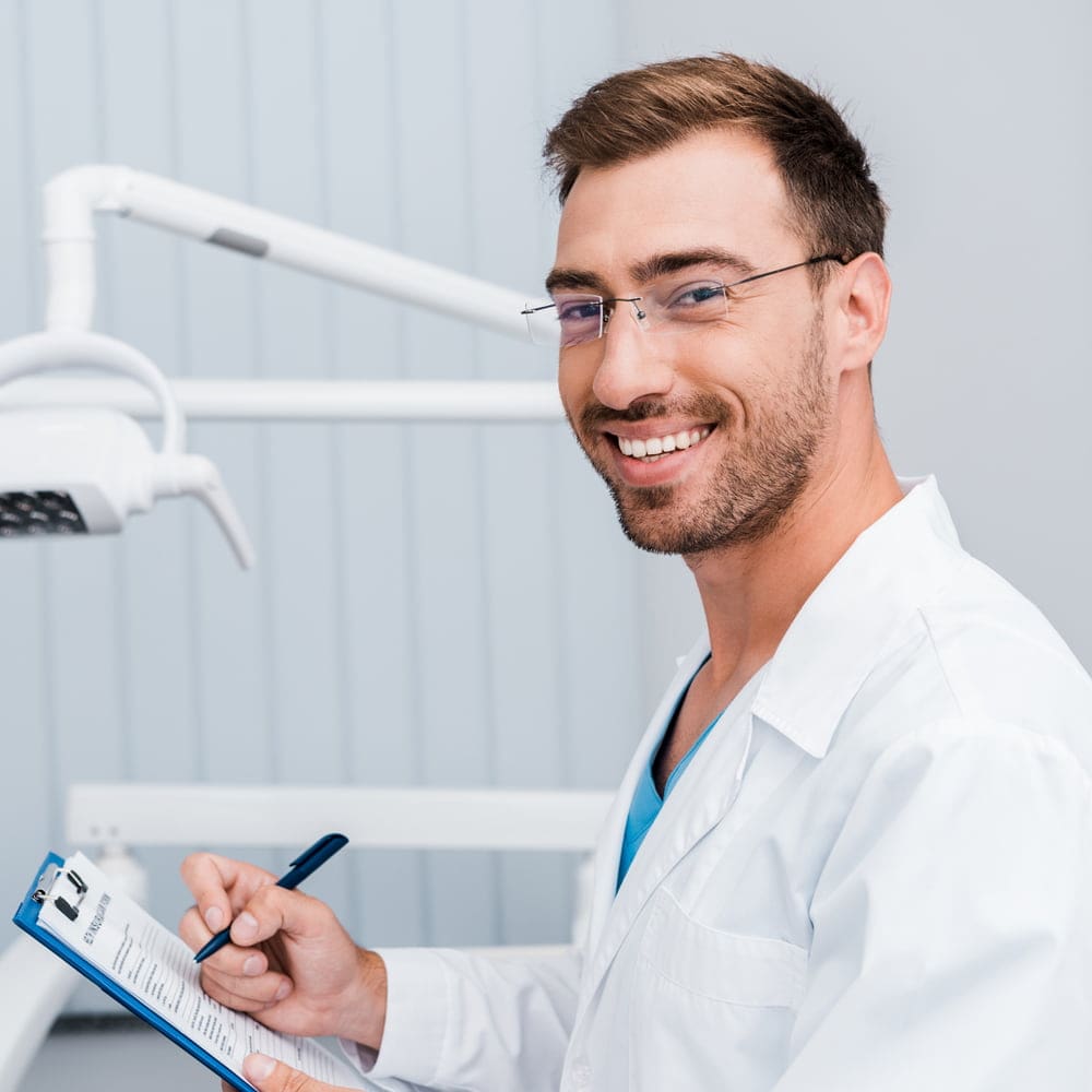 A male dentist in a white coat smiles while holding a clipboard and pen in a dental office in Austin