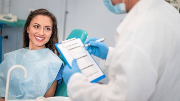 A woman sits in a dental chair, smiling at a dentist who is holding a clipboard.