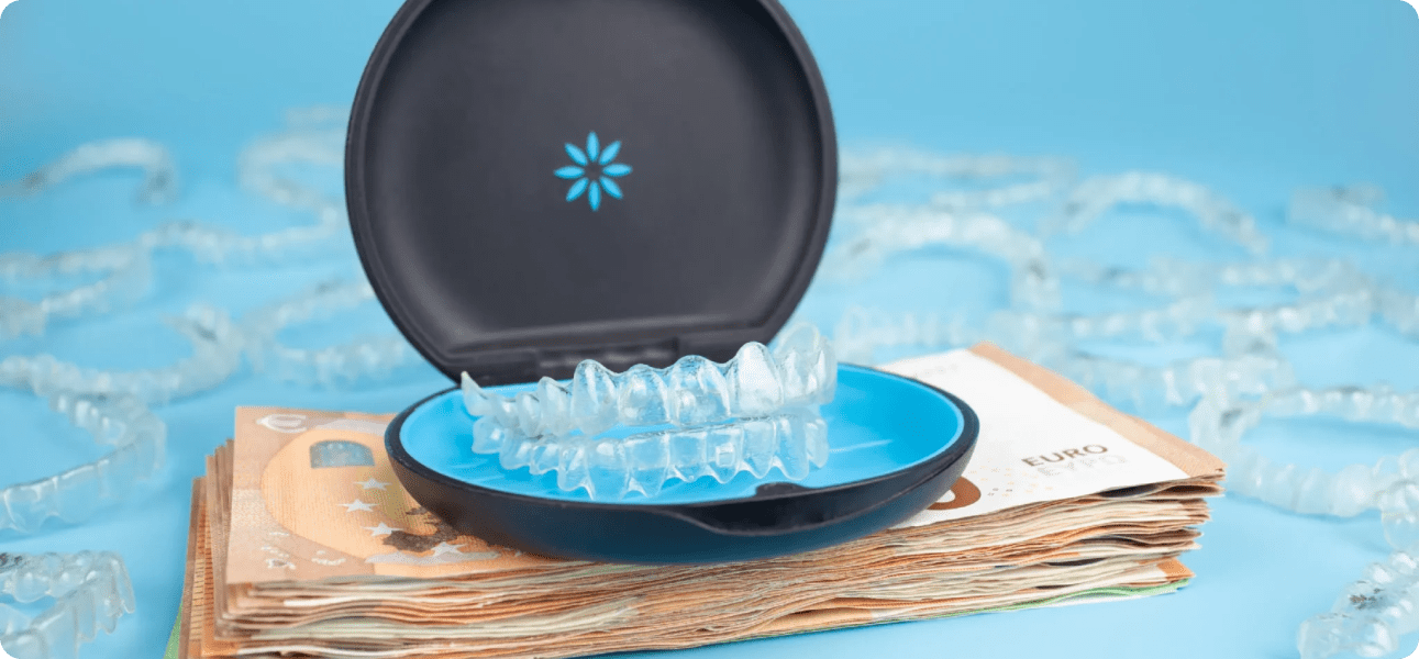 A dental aligner case is open, displaying clear aligners. It is placed on a stack of Euro banknotes with several aligners scattered in the background against a blue backdrop.