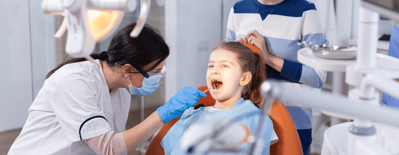 A dentist examines a young girl's teeth in a dental clinic. The girl is seated in an orange dental chair and another person stands nearby. Dental equipment is visible.