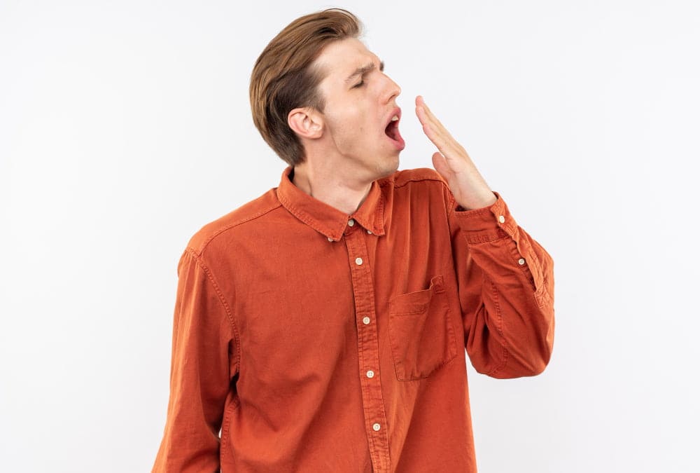 A person wearing an orange shirt yawns with their hand near their mouth against a plain white background.