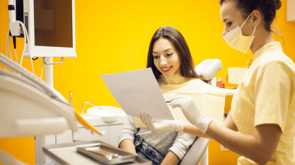 A dental professional shows a female patient a document in a brightly lit dental office with orange walls. The patient sits in a dental chair.