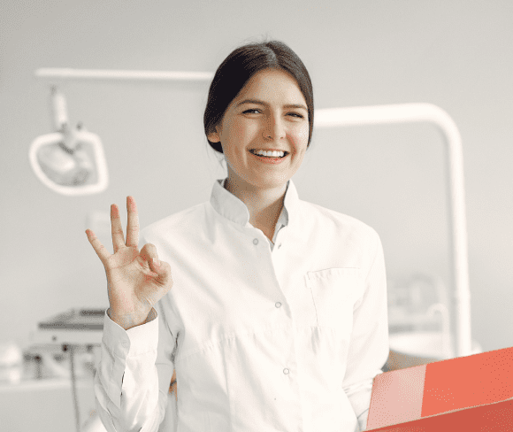 A woman in a white coat stands in a dental office, smiling and making an "OK" hand gesture.