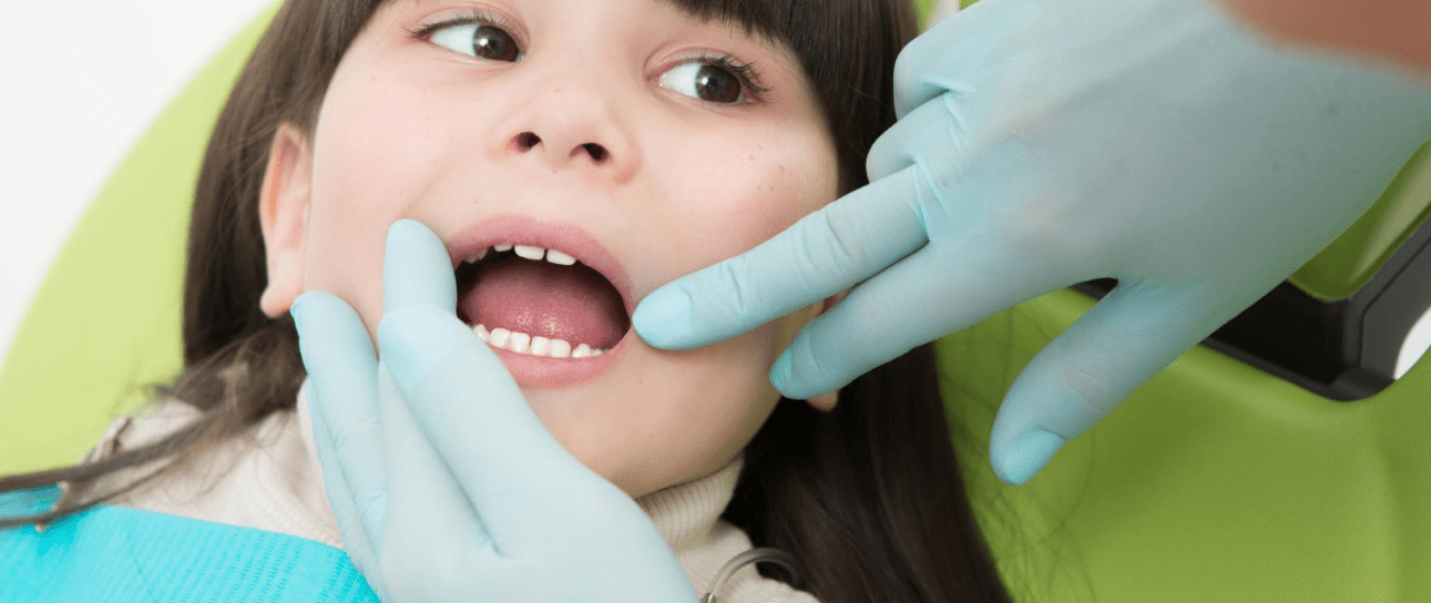 A child in a dental chair with a dentist examining their teeth, wearing blue gloves.