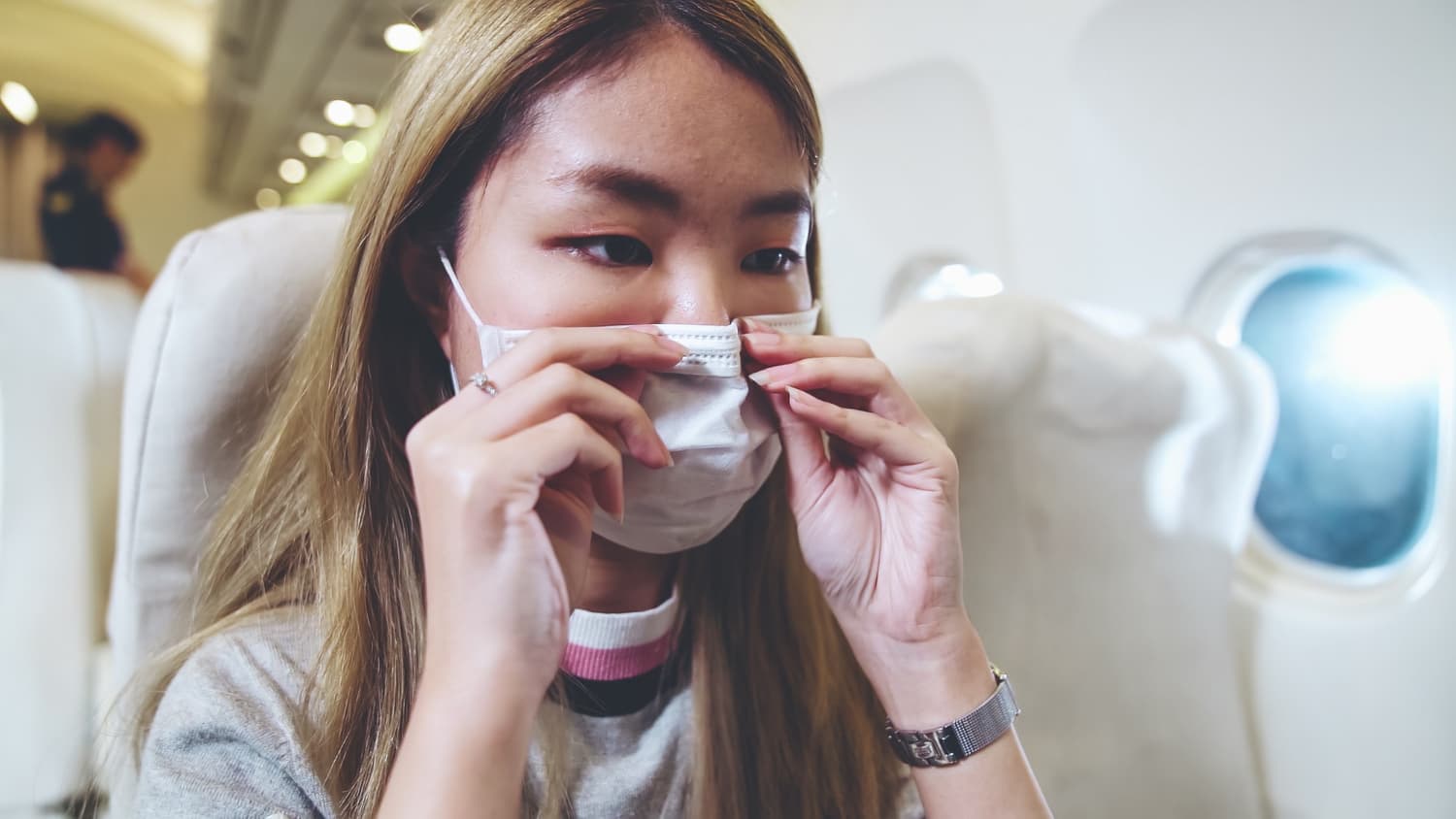 A person sitting in an airplane adjusts their face mask while looking out the window