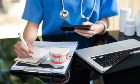A healthcare professional in blue scrubs writes in a notebook and holds a phone. A dental model, laptop and medicine bottles are on the desk.