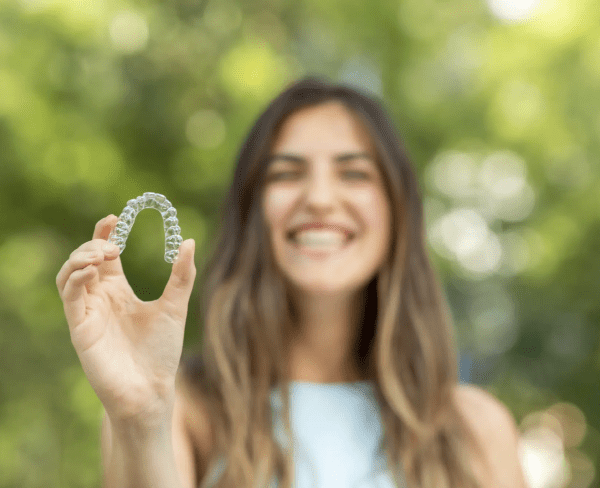 Person holding a clear orthodontic aligner with a blurred smile in the background and greenery around.
