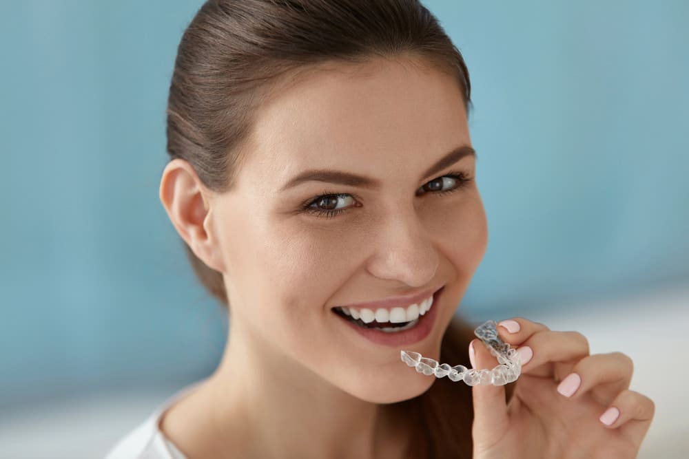 A woman smiles while holding a clear dental aligner near her mouth against a blue background, showcasing the results of the best orthodontics.