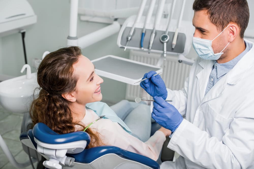 Dentist in a mask and gloves examines a smiling patient in a dental chair with instruments in hand.