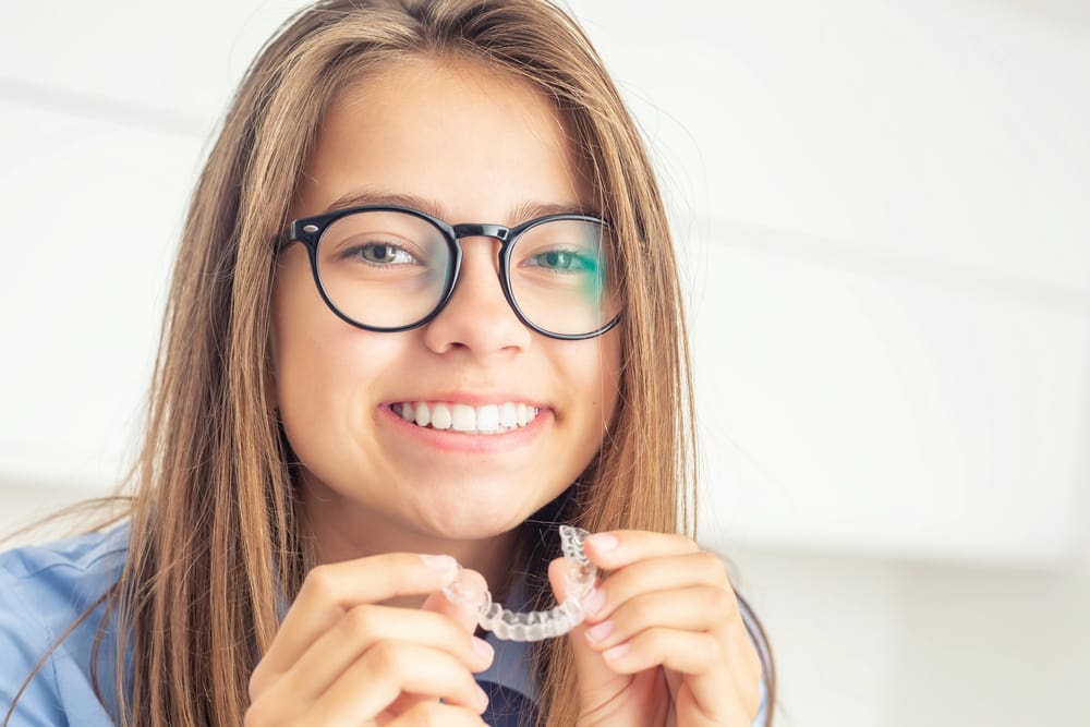 A smiling person with glasses holds a clear dental retainer in their hands.