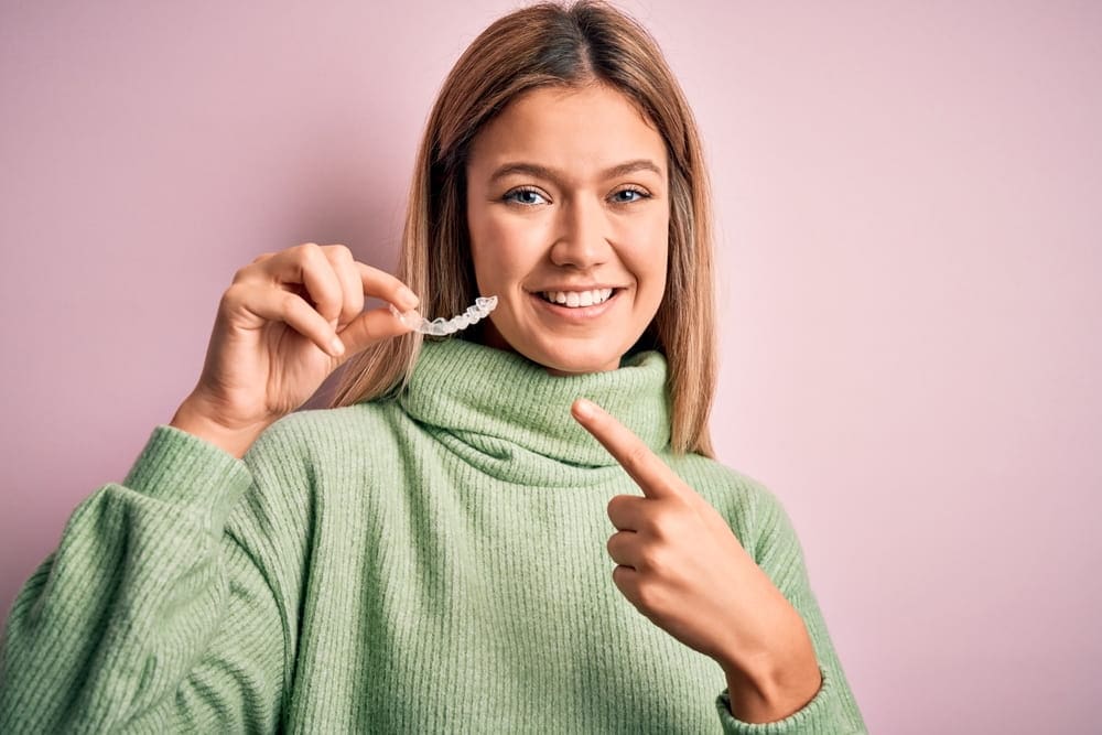 Woman in a green sweater holding and pointing at a clear dental aligner against a pink background.