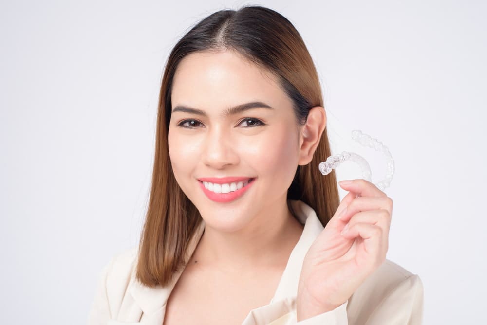 A woman smiling and holding a clear dental aligner near her face, wearing a cream-colored jacket, with a plain white background.