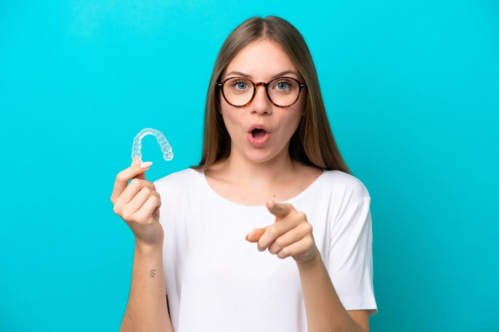 Woman in glasses holding a clear dental aligner against a blue background, pointing forward.