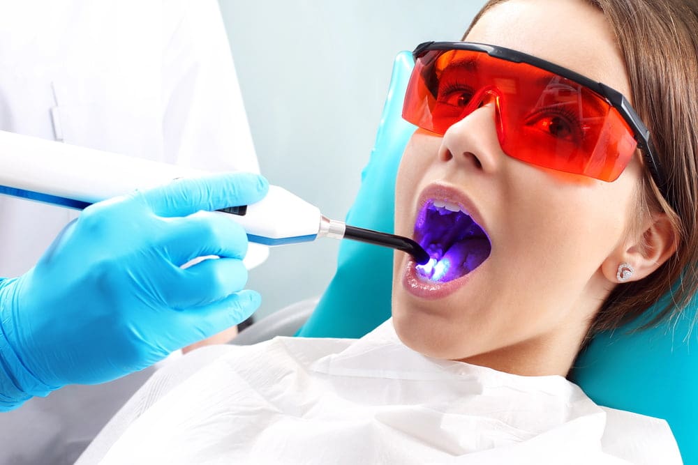A dentist uses a curing light on a patient's teeth. The patient wears protective red glasses and a dental bib