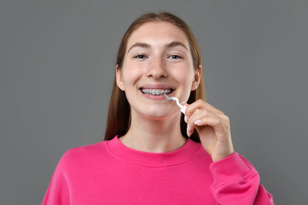 Young person with long hair in a pink sweater cleaning braces with a dental tool against a gray background