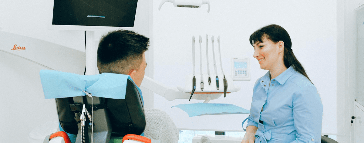 A dentist in a blue coat talks to a seated patient in a dental office with equipment visible in the background.