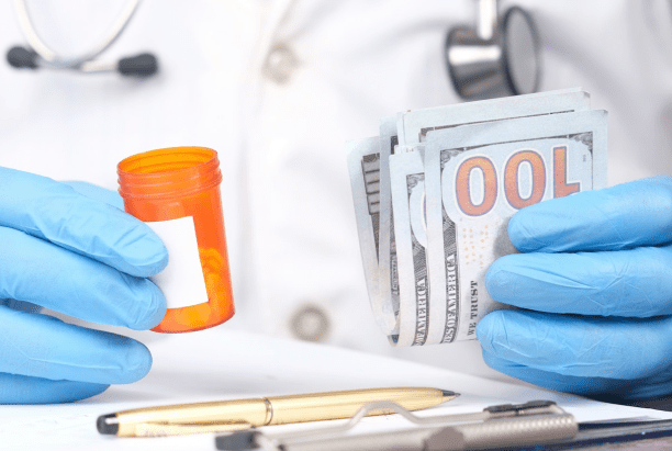 A person in a lab coat and blue gloves holds a small pill bottle in one hand and a stack of hundred-dollar bills in the other, with a pen and clipboard visible on a desk.
