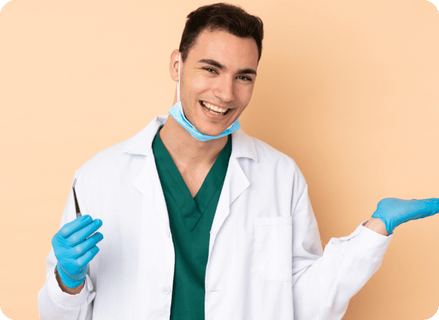 A person in a white coat and green scrubs smiles, holding a dental tool in one hand and gesturing with the other. They wear blue gloves and a face mask under their chin.