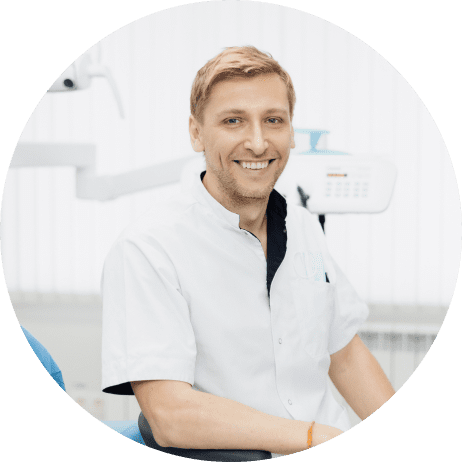 A dentist in a white coat smiles while seated in a dental office with equipment in the background.