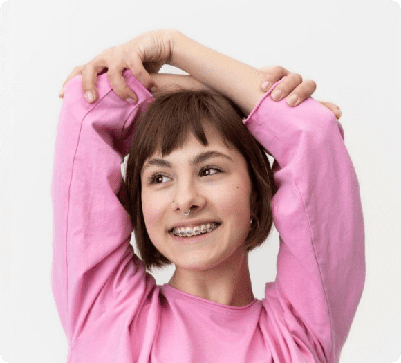 A person with short brown hair, braces and a septum piercing smiles while stretching their arms above their head, wearing a pink long-sleeve shirt.