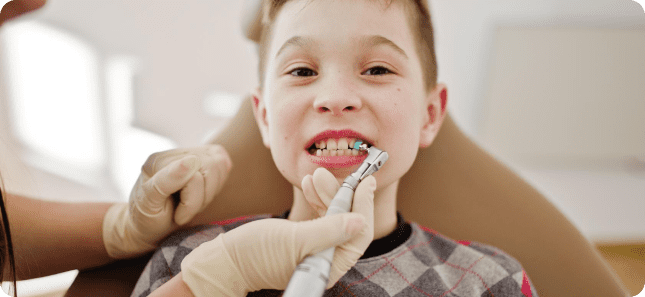 A child sits in a dental chair while a dentist, wearing gloves, cleans their teeth with a dental tool.