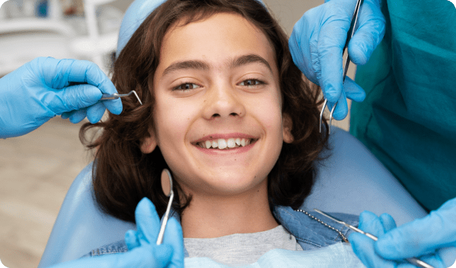 Child smiling in dental chair with dentist's hands and tools around mouth.