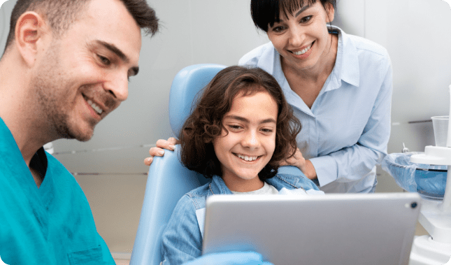 A dentist and a woman smile at a girl sitting in a dental chair, who is holding a tablet.