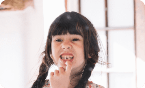 Child with dark hair and braided pigtails smiles, showing a gap where a tooth is missing, in a brightly lit room.