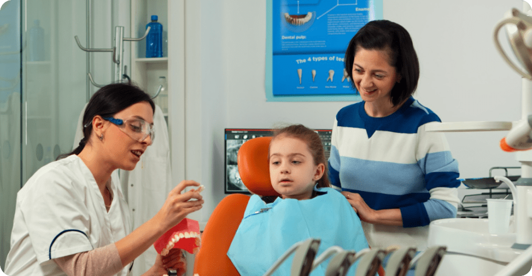A dentist shows a dental model to a young girl sitting in a chair, while an adult stands beside them.