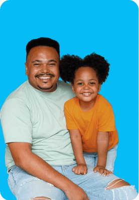 A man in a light green shirt and ripped jeans sits with a smiling child in an orange shirt against a blue background.