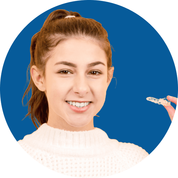 A person with long hair in a ponytail holds a clear dental aligner while smiling. The background is a solid blue.