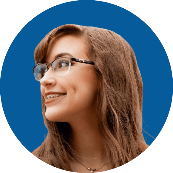 A person with long brown hair, glasses and braces smiles while looking to the side against a blue background.
